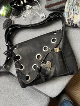 Cartera de cuero negra con correa regulable, estilo bandolera o al hombro. Decorada con charm de corazón metálico, encaje negro, cadenas y aros metálicos.