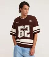 Remera oversized deportiva color marrón con cuello en V, rayas blancas en las mangas y en la parte inferior, y el número "62" estampado en blanco en el frente. Incluye el bordado "Humanity" y un logo de globo terráqueo.