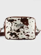 Morral de cuero marca Tranquera con frente de pelo natural en diseño animal print de vaca. Presenta una estructura rectangular con bordes redondeados, correa de cuero regulable y desmontable mediante mosquetones metálicos, y una placa con el logo de la marca en el centro.
