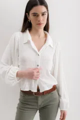 Camisa blanca con textura arrugada, corte cropped, cuello camisero, mangas largas abullonadas y cierre frontal con botones.