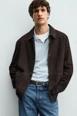 Sobrecamisa marrón de cuero gamuzado, corte recto, con cuello clásico, cierre frontal con botones, mangas largas con puños abotonados y bolsillos de parche en el pecho.
