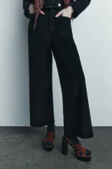 Pantalón ancho de pana color negro, de tiro medio, con trabillas en la cintura, bolsillos delanteros y cierre frontal con cremallera, botón y gancho metálico.