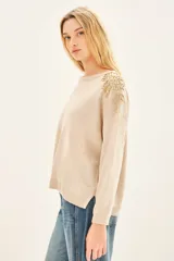 Sweater color beige con cuello redondo y mangas largas. Presenta apliques dorados en los hombros y un cinturón marrón con forma de flor.