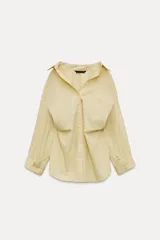 Camisa oversize color amarillo claro a rayas finas verticales azules, de cuello solapa y manga larga tipo murciélago acabada en puño con hombro caído. Tiene un bolsillo de plastrón en el delantero y cierre frontal con botones.