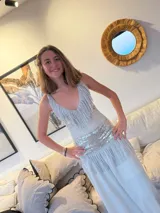 Vestido largo blanco con escote en V, decorado con flecos cortos en el busto y la falda. Presenta una franja de lentejuelas plateadas en el busto y la cadera. La espalda queda descubierta, con breteles metalizados que se cruzan en la espalda.