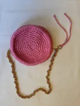 Cartera tejida a crochet color rosa, con forma circular y apliques bordados de labios, plumas, planetas y una mano. Tiene una correa de cadena dorada entrelazada con trapillo rosa, cierre superior con cremallera y forro interior.