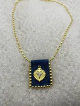 Collar con cadena dorada y dije rectangular de tela de jean con borde de pequeñas bolitas doradas. El dije tiene un aplique dorado con forma de corazón con una cruz en la parte superior y strass incrustados.