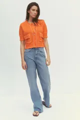 Chaqueta naranja de punto con cuello redondo y manga corta. Presenta bolsillos delanteros de parche con solapa y cierre frontal con botones. Detalle de pespuntes blancos a contraste.