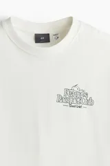 Remera blanca de corte holgado con cuello redondo y estampado de Snoopy con la leyenda "Peanuts Racquet Club" en el pecho.