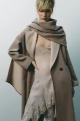Abrigo largo color beige confeccionado en lana. Presenta cuello solapa con bufanda desmontable, manga larga, bolsillos laterales y cierre frontal cruzado con botones.