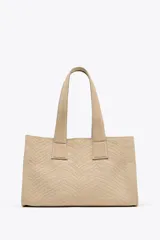 Bolso shopper color beige con diseño trenzado y doble asa de hombro. Cuenta con un bolsillo interior con cierre de cremallera y cierre principal con clip.