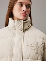 Campera puffer color beige claro con logo de Calvin Klein bordado en el pecho. Tiene cuello alto, cierre con cremallera y botones a presión, bolsillos laterales y puños elásticos.