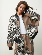 Campera bomber con estampado de flores en blanco y negro sobre fondo beige y marrón. Tiene cuello, puños y bajo acanalados en negro, y cierre frontal con cremallera.