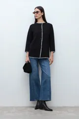 Saco negro de tejido de punto, con cuello redondo y cierre frontal de un solo botón. Presenta un ribete de costura contrastante en color blanco a lo largo de todo el borde, incluyendo puños y bolsillos.