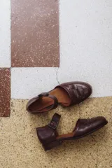 Zapato marrón de cuero con correa al tobillo y hebilla negra. Diseño trenzado en la parte delantera.