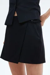 Short pollera negro de tiro alto con diseño cruzado tipo pareo y cinturón fijo con hebilla revestida. Presenta cierre oculto y cierre de corchete en un lateral.