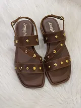 Sandalias bajas de cuero marrón con tiras cruzadas y apliques de tachas doradas.