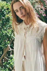 Blusa sin mangas color blanco con cuello alto, bordados y puntillas combinadas a tono, y apliques de abalorios. Cierre frontal con abertura, lazada y botón.