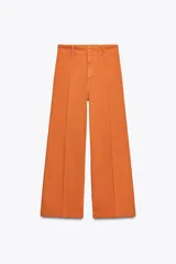 Pantalón de jean color naranja, de tiro alto y pierna ancha. Confeccionado en hilatura con mezcla de algodón. Presenta trabillas en la cintura, bolsillos de plastrón en la espalda y costura marcada en el delantero. Cierre frontal con cremallera, botón y gancho metálico.