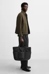 Bolso tipo shopper negro fabricado en tejido técnico. Cuenta con un compartimento principal con cierre de cremallera y un bolsillo interior con cierre. En la parte frontal tiene tres bolsillos. Tiene dos asas de mano.