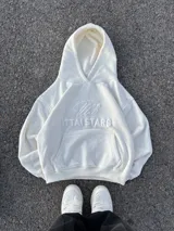 Hoodie color blanco roto (off-white) con capucha y bolsillo canguro. Presenta un bordado en relieve en el pecho con un diseño de caligrafía y el texto "ITTAI STARS" debajo.