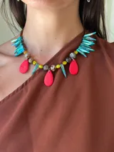 Collar con colgantes asimétricos, compuesto por cuentas de cristal checo, piedras facetadas y colgantes en forma de lágrima de color rojo vibrante. Presenta detalles en forma de colmillos de nácar teñidos de color turquesa iridiscente.