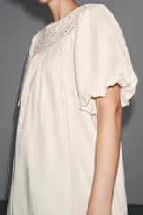 Vestido corto color crudo confeccionado en hilatura con mezcla de algodón y lino. Presenta cuello redondo, manga corta abullonada, detalle de bordados a tono y bajo recto. Cierre en espalda con abertura y botón.