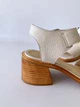 Sandalias de cuero color crudo, con tira ancha sobre los dedos, pulsera al tobillo con hebilla metálica y taco de madera de 6 cm de altura.