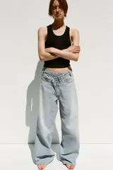 Pantalón de jean celeste de tiro alto, corte wide leg y largo hasta el tobillo. Presenta un diseño asimétrico en la cintura con cierre frontal cruzado con cremallera y botones metálicos.