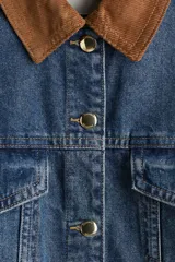 Campera de denim azul con cuello de pana marrón, botones metálicos dorados, canesú delantero y trasero, corte holgado, hombros caídos, puños abotonados, bolsillos laterales discretos y bolsillos superiores con solapa y botón.