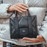 Cartera chica negra de símil cuero soft, con acceso por cierre, correa regulable y asas fijas. Tiene un bolsillo interno con cierre y otro abierto, herrajes plateados y logotipo XL Extra Large en el frente.