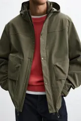 Parka verde militar confeccionada en tejido técnico ligero resistente al viento, con cuello alto, capucha, manga larga con puños ajustables, bolsillos de plastrón con solapa en la cadera, bolsillo interior, bajo ajustable con elásticos y cierre frontal con cremallera oculta por solapa con botones a presión.