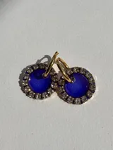 Par de aros colgantes con forma circular, de resina color azul y borde con strass. El arito es de acero quirúrgico color dorado.