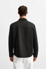Sobrecamisa negra de tejido con elasticidad, cuello solapa y manga larga acabada en puño con botón a presión. Tiene bolsillos de plastrón en el pecho y cierre frontal con botones a presión.