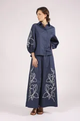 Pantalón ancho de algodón color celeste claro, con bordados de flores blancas en los bajos. Presenta un corte de tiro alto con pliegues frontales y cierre de botón visible.