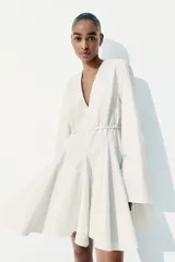 Vestido corto blanco con escote en V profundo, mangas largas con abertura en los puños, cierre oculto en la espalda y cinturón fino extraíble.
