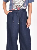 Pantalón de jean azul oscuro de tiro alto, corte ancho y pierna acampanada. Presenta pliegues frontales y un lazo de tela a tono para ajustar en la cintura.