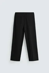 Pantalón negro de corte ancho con cintura elástica, pliegues frontales, bolsillos laterales y traseros de vivo. Tiene detalle de hebilla metálica en la trabilla y cierre frontal con cremallera y botón.