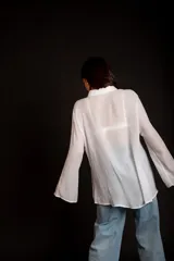 Camisa color beige claro, de corte holgado, con cuello clásico, cierre frontal con botones, mangas largas con puños abotonados y detalles de broderie en la parte delantera.