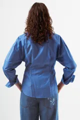 Camisa de manga larga color azul con finas rayas verticales blancas. Presenta cuello mao, botones frontales y dos bolsillos de parche con tapa en el pecho. La prenda tiene un diseño ajustado en la cintura con pinzas y un volumen holgado en las mangas.