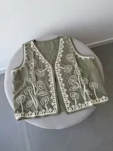 Chaleco sin mangas color beige claro, con bordados florales en relieve y abertura frontal.