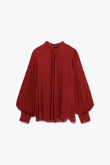 Blusa de cuello subido color ladrillo, confeccionada en hilatura de viscosa 100%. Manga larga con pliegues y abullonada. Detalle aplicación de lazo en cuello ajustable con lazada. Cierre frontal con botones.
