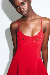 Vestido corto rojo, ajustado en el busto y con falda acampanada. Tiene tirantes finos ajustables y apliques metálicos dorados en forma de corazón y redondos en el escote.