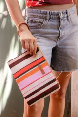 Sobre de algodón a rayas verticales de colores naranja, rosa, marrón, rojo y blanco. Tiene cierre superior con cremallera y tira de cuero marrón para la muñeca.