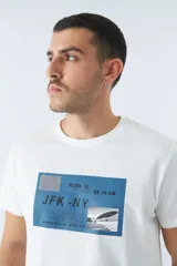 Remera blanca de algodón con cuello redondo y mangas cortas. Estampado frontal con diseño rectangular en tonos azules que incluye texto, coordenadas y la imagen de un avión.