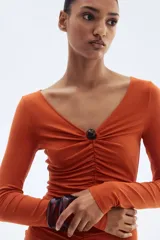 Vestido mini color naranja, entallado, de punto elástico con leve brillo, manga larga, escote en V, cuenta decorativa en el centro del busto y costura fruncida adelante para un efecto drapeado.