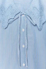Camisa celeste a rayas verticales blancas, de corte recto, con cuello bobo con bordados y escote pico con lazada. Tiene mangas cortas abullonadas con elástico y cierre frontal con botones.