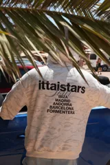 Buzo canguro con capucha, color blanco con estampado de camuflaje en tonos grises. Estampado en la espalda con el logo de la marca y nombres de ciudades.