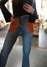 Pantalón de jean celeste con cintura elastizada, corte recto, bolsillos laterales y aplique de cuero marrón en el bolsillo delantero. Presenta botones metálicos en los laterales de la cintura y etiqueta de cuero en la parte trasera.