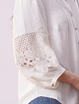 Blusa blanca de gasa con cuello a la base, escote en V y abertura central con botones. Tiene mangas 3/4 amplias con apliques de crochet y detalles plisados en la parte delantera.